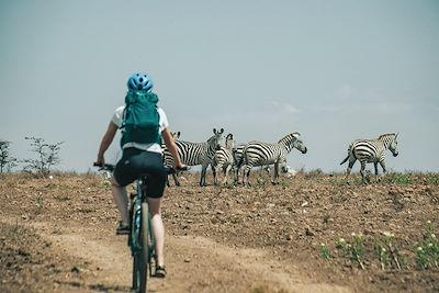 © Hillary Matemu - Femme faisant du vélo à côté des zèbres - Région Kilimandjaro - Tanzanie Femme faisant du vélo à côté des zèbres - Région Kilimandjaro - Tanzanie